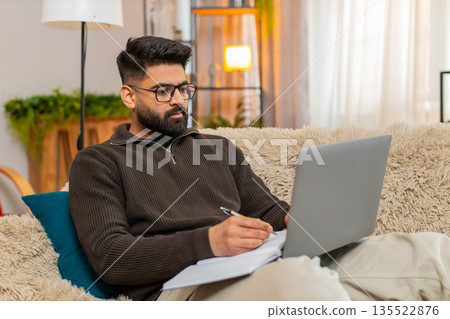 Indian man writing notes from laptop into notebook at home on sofa with focused look studying online 135522876