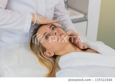Young woman facial skin check by cosmetologist in gloves before planned relaxing cosmetic procedure Young woman facial skin check by cosmetologist in gloves before planned relaxing cosmetic procedure 135522888