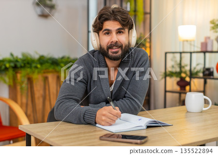 Indian man listens to audiobook on headphones takes notes in notebook focused intense study session 135522894