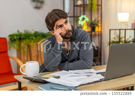 Indian man holds head over unpaid bills on table with calculator and laptop worried about money debt 135522895
