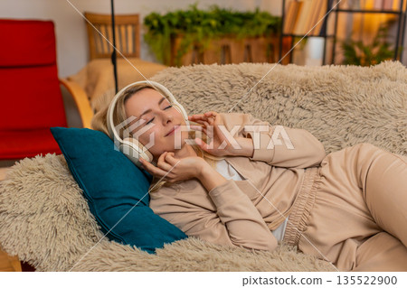 Woman lying on couch with eyes closed, listening to music through headphones in peaceful relaxation 135522900