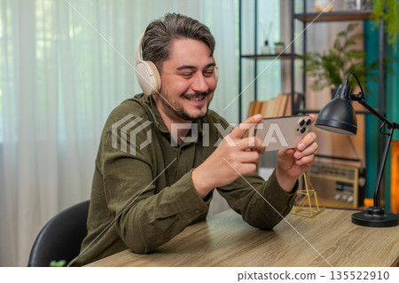 Middle-aged man bursts laughing to comedy audio smartphone headphones shaking head joyful at table 135522910