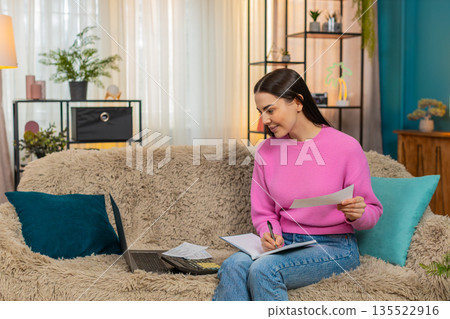 Young woman on sofa with laptop and calculator counting expenses worried writing notes in notebook 135522916