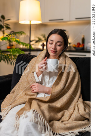 Young woman drinks hot tea slow sips warms hands in blanket gentle smile calm break restful evening Young woman drinks hot tea slow sips warms hands in blanket gentle smile calm break restful evening 135522949