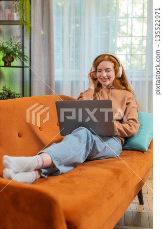 Happy relaxed Caucasian young woman in wireless headphones listening music using laptop on sofa 135522971