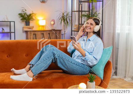 Joyful young woman enjoying listening to music through wireless headphones dancing at home room sofa 135522973