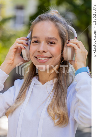 Happy young woman taking off headphones after listening favorite energetic music on city street Happy young woman taking off headphones after listening favorite energetic music on city street 135523378