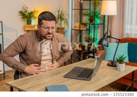 Disappointed freelancer man feeling stomach ache overeating during work process on laptop at home Disappointed freelancer man feeling stomach ache overeating during work process on laptop at home 135523380