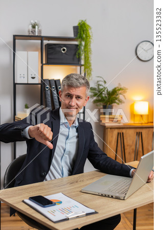 Upset young Caucasian freelancer in suit using laptop and showing thumb down at home office table Upset young Caucasian freelancer in suit using laptop and showing thumb down at home office table 135523382