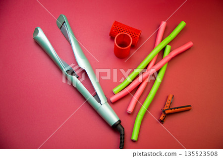 Top view of a green hair straightener with a twisted ceramic canvas, next to which there are curlers of different shapes and sizes on a pink background. The concept of styling, hairstyle, curls. High 135523508