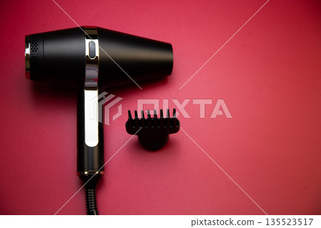 Close-up of a black hair dryer with a comb attachment to create volume on a pink background with space for text. The concept of long-term hair styling, hairstyles, curls and hair care. High quality Close-up of a black hair dryer with a comb attachment to create volume on a pink background with space for text. The concept of long-term hair styling, hairstyles, curls and hair care. High quality 135523517