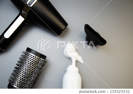 Close-up of hair styling foam in a white bottle, next to it a hairdryer with a round comb to create voluminous hairstyles on a gray background with space for text. The concept of long-term hair Close-up of hair styling foam in a white bottle, next to it a hairdryer with a round comb to create voluminous hairstyles on a gray background with space for text. The concept of long-term hair 135523532