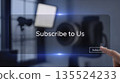 Digital screen shows message to subscribe, hand gestures towards button in room with camera equipment and soft lights during evening hours 135524233