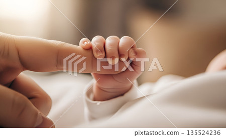 Small hand holds adult finger in a warm indoor setting during a quiet moment of connection between a parent and baby 135524236