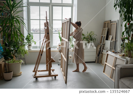 Focused woman painter artist finishing painting and taking canvas from easel at home workshop. 135524667