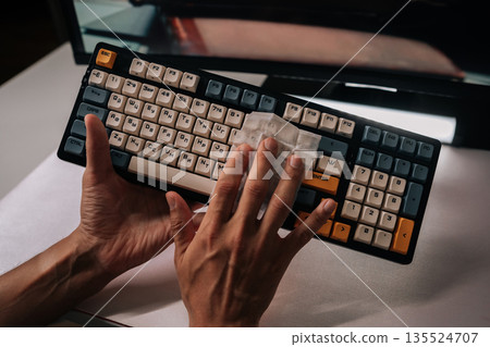 Man cleaning mechanical keyboard with disposable wipe, maintaining hygiene, ensuring germ-free surfaces, performing regular maintenance on office equipment to keep functional and clean. Man cleaning mechanical keyboard with disposable wipe, maintaining hygiene, ensuring germ-free surfaces, performing regular maintenance on office equipment to keep functional and clean. 135524707