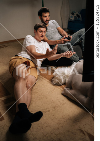 Vertical shot of two friends bonding and enjoying leisure time at home, deeply engaged in competitive console gaming session, using wireless controllers for interactive entertainment and digital fun. Vertical shot of two friends bonding and enjoying leisure time at home, deeply engaged in competitive console gaming session, using wireless controllers for interactive entertainment and digital fun. 135524715