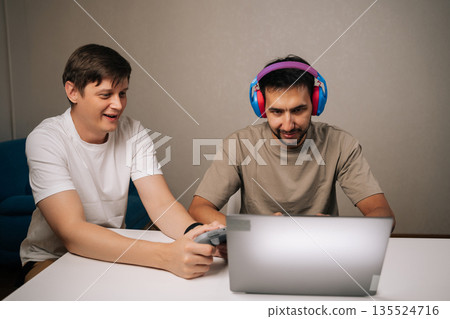 Relaxed two male friends enjoying competitive video gaming on laptop sitting at desk at home, connecting and having fun listening with headphones and concentrating using game controller. Relaxed two male friends enjoying competitive video gaming on laptop sitting at desk at home, connecting and having fun listening with headphones and concentrating using game controller. 135524716