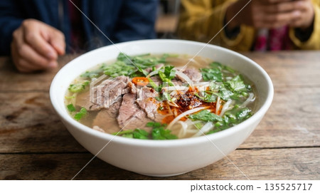 Two people are sitting at a wooden table and enjoying bowls of noodle soup filled with meat and herbs. They are in an Asian restaurant during lunch time focusing on their meal. 135525717