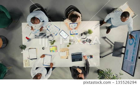Top down view of corporate employees gathered in a meeting to discuss finance, objectives and proposals. Colleagues share insights, documents and metrics to guide project planning. 135526294