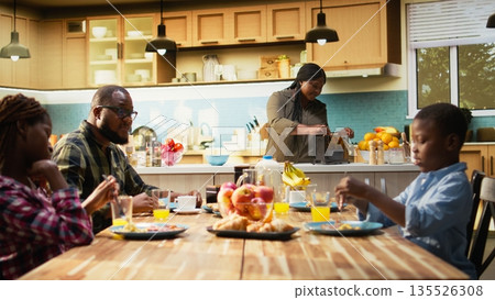 African American mother preparing breakfast in the morning light while the family wait at the table with croissants, fruits and orange juice. Parents and children laugh together, love. Camera A. African American mother preparing breakfast in the morning light while the family wait at the table with croissants, fruits and orange juice. Parents and children laugh together, love. Camera A. 135526308