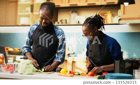 Black young people cutting fresh vegetables and adding in a bowl to make delicious salad, having carrots, bell peppers and cucumbers on cutting board. Vegetarian people preparing lunch. Black young people cutting fresh vegetables and adding in a bowl to make delicious salad, having carrots, bell peppers and cucumbers on cutting board. Vegetarian people preparing lunch. 135526315