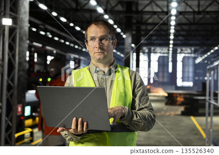 Supervisor inspecting steel equipment with laptop and safety glasses on the factory floor, reflecting industrial manufacturing process expertise and manpower supporting heavy production. Supervisor inspecting steel equipment with laptop and safety glasses on the factory floor, reflecting industrial manufacturing process expertise and manpower supporting heavy production. 135526340