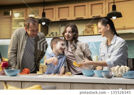 Family time in the kitchen during pizza night with parents and kids, adding toppings like mozzarella and pepperoni. Having fun with tasty homemade food, surrounded by laughter and joy. 135526345