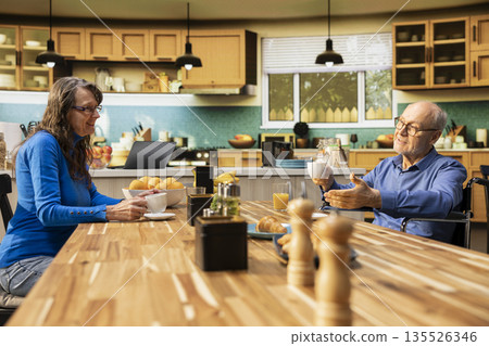 Mature man with physical disability serving coffee and pastry with his wife, enjoying cozy morning routine with laughter and casual conversation. Positive household moment in their modern home. Mature man with physical disability serving coffee and pastry with his wife, enjoying cozy morning routine with laughter and casual conversation. Positive household moment in their modern home. 135526346