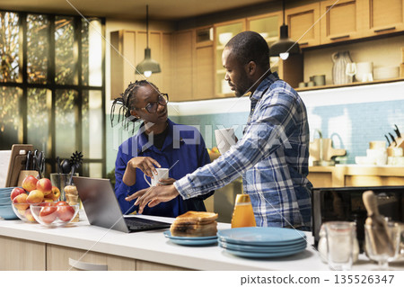 Happy couple preparing a delicious breakfast together and drinking coffee, following an online recipe on laptop. Getting ready to eat toast slices and homemade pastry, healthy food in the kitchen. Happy couple preparing a delicious breakfast together and drinking coffee, following an online recipe on laptop. Getting ready to eat toast slices and homemade pastry, healthy food in the kitchen. 135526347