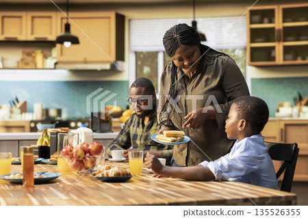 Parents and children gathered in the kitchen for breakfast and serving coffee, cereal and homemade food. Morning light fills the room as family shares laughter and a peaceful home routine. Parents and children gathered in the kitchen for breakfast and serving coffee, cereal and homemade food. Morning light fills the room as family shares laughter and a peaceful home routine. 135526355