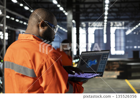 Black supervisor inspecting turbine production on laptop software in industry plant, monitoring heavy equipment performance. Warehouse features metal production tools and machinery. 135526356