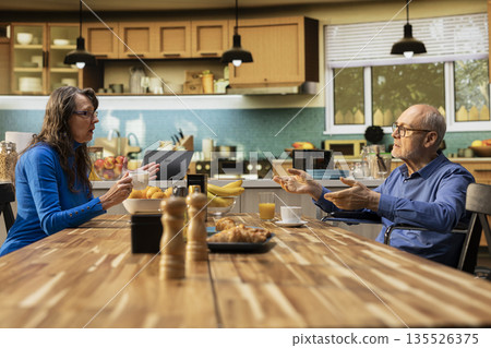 Husband in wheelchair and his wife serving coffee and chatting at the breakfast table, candid moment reflecting healthy lifestyle and comfort in retirement. Elderly people in their home kitchen. 135526375