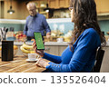 Mockup display on phone at kitchen table with elderly woman smiling and relaxing, serving coffee and meal for breakfast at the table. Fun retirement lifestyle from comfortable rustic home. 135526404