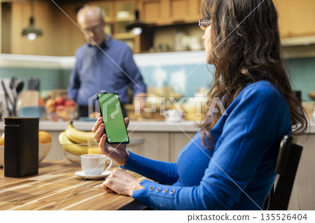 Mockup display on phone at kitchen table with elderly woman smiling and relaxing, serving coffee and meal for breakfast at the table. Fun retirement lifestyle from comfortable rustic home. Mockup display on phone at kitchen table with elderly woman smiling and relaxing, serving coffee and meal for breakfast at the table. Fun retirement lifestyle from comfortable rustic home. 135526404