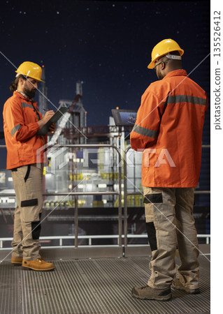 Offshore platform crew using tablet to oversee drilling operations during night shift. Drilling barge staff members reviewing sensor data on device to ensure continuous production 135526412