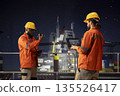 Offshore rig employee uses laptop on oil platform deck next to colleague using walkie talkie late at night. Barge engineer reviewing energy operations on drilling rig next to african american coworker 135526417