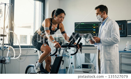 athlete is riding a stationary bike in a lab. A medical professional monitors the athlete's performance and health data on a tablet. Several sensors are attached to the athlete's body. athlete is riding a stationary bike in a lab. A medical professional monitors the athlete's performance and health data on a tablet. Several sensors are attached to the athlete's body. 135527079