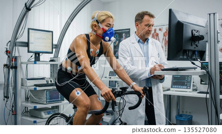 athlete is riding a stationary bike while wearing a mask connected to equipment. A coach monitors the data on a computer and records results. athlete is riding a stationary bike while wearing a mask connected to equipment. A coach monitors the data on a computer and records results. 135527080