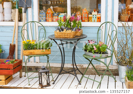 Cozy spring flower shop exterior with potted plants, seedlings in boxes, rustic chairs and table Cozy spring flower shop exterior with potted plants, seedlings in boxes, rustic chairs and table 135527157