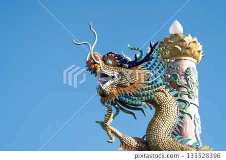 Ornate Chinese dragon sculpture with golden scales and vibrant mane against a clear blue sky. Traditional architectural element from a temple, showcasing intricate craftsmanship and heritage. 135528396