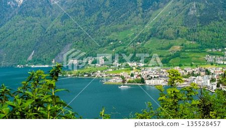 Enchanting Settlement Along Riverbank Surrounded By Vibrant Flora And Impressive Mountain Ranges 135528457