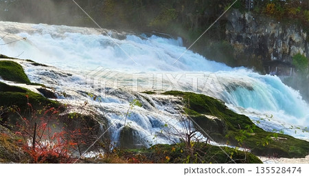 Vivid Natural Landscape Featuring Cascading Waterfall Surrounded By Vibrant Vegetation And Rocky Formations 135528474