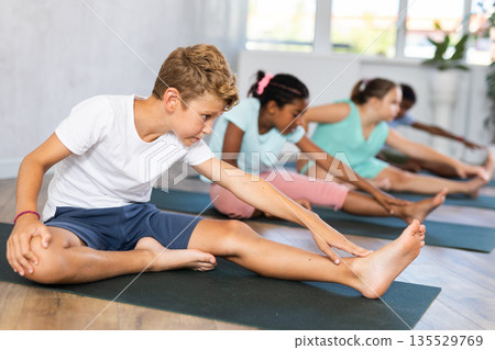 Kids practicing yoga, doing Head to Knee Forward Bend exercise, Janu Sirsasana pose, working out in yoga studio. 135529769