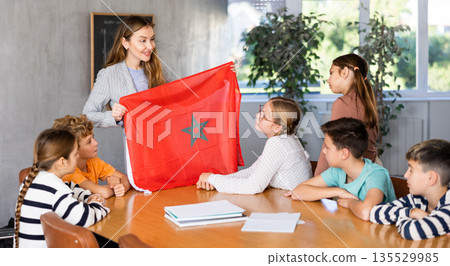 Smiling young female teacher conducting lesson for preteens, showing state flag of Morocco in audience of middle school Smiling young female teacher conducting lesson for preteens, showing state flag of Morocco in audience of middle school 135529985