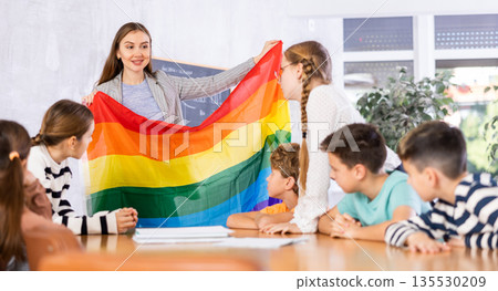 Young female teacher showing flag of LGBT to schoolchildren preteens and explaining LGBTQ social movements in classroom Young female teacher showing flag of LGBT to schoolchildren preteens and explaining LGBTQ social movements in classroom 135530209
