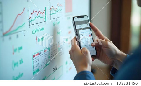 A suited individual studies a smartphone displaying financial graphs, emphasizing data-driven decision-making in a tech-savvy environment A suited individual studies a smartphone displaying financial graphs, emphasizing data-driven decision-making in a tech-savvy environment 135531450