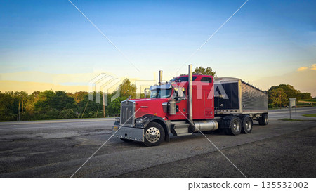 bold red semi truck with a long trailer sits in an open lot during sunset. The powerful rig highlights industrial design, chrome accents, and the rugged appeal of transportation and logistics 135532002