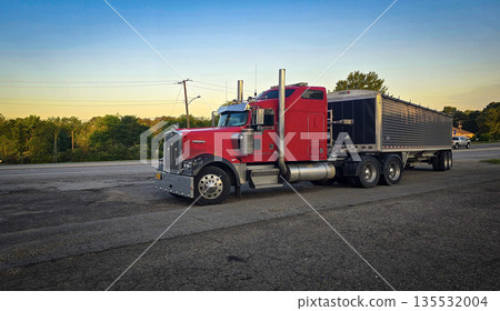 bold red semi truck with a long trailer sits in an open lot during sunset. The powerful rig highlights industrial design, chrome accents, and the rugged appeal of transportation and logistics 135532004