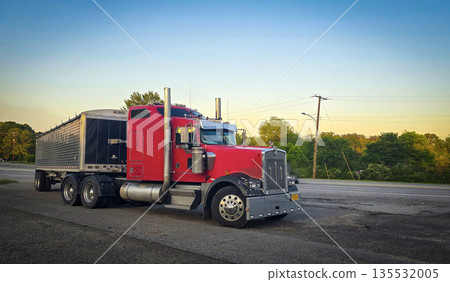 bold red semi truck with a long trailer sits in an open lot during sunset. The powerful rig highlights industrial design, chrome accents, and the rugged appeal of transportation and logistics 135532005